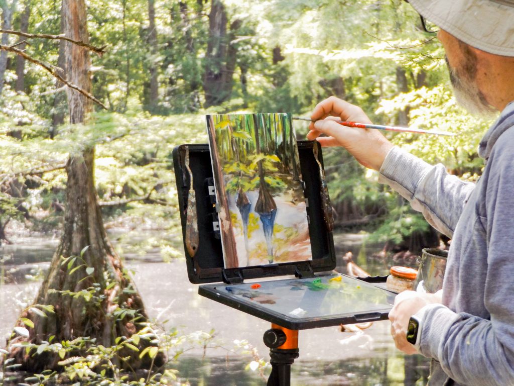 Man is painting in oil pants while looking outward to a swamp filled with cypress trees on a plein air palette