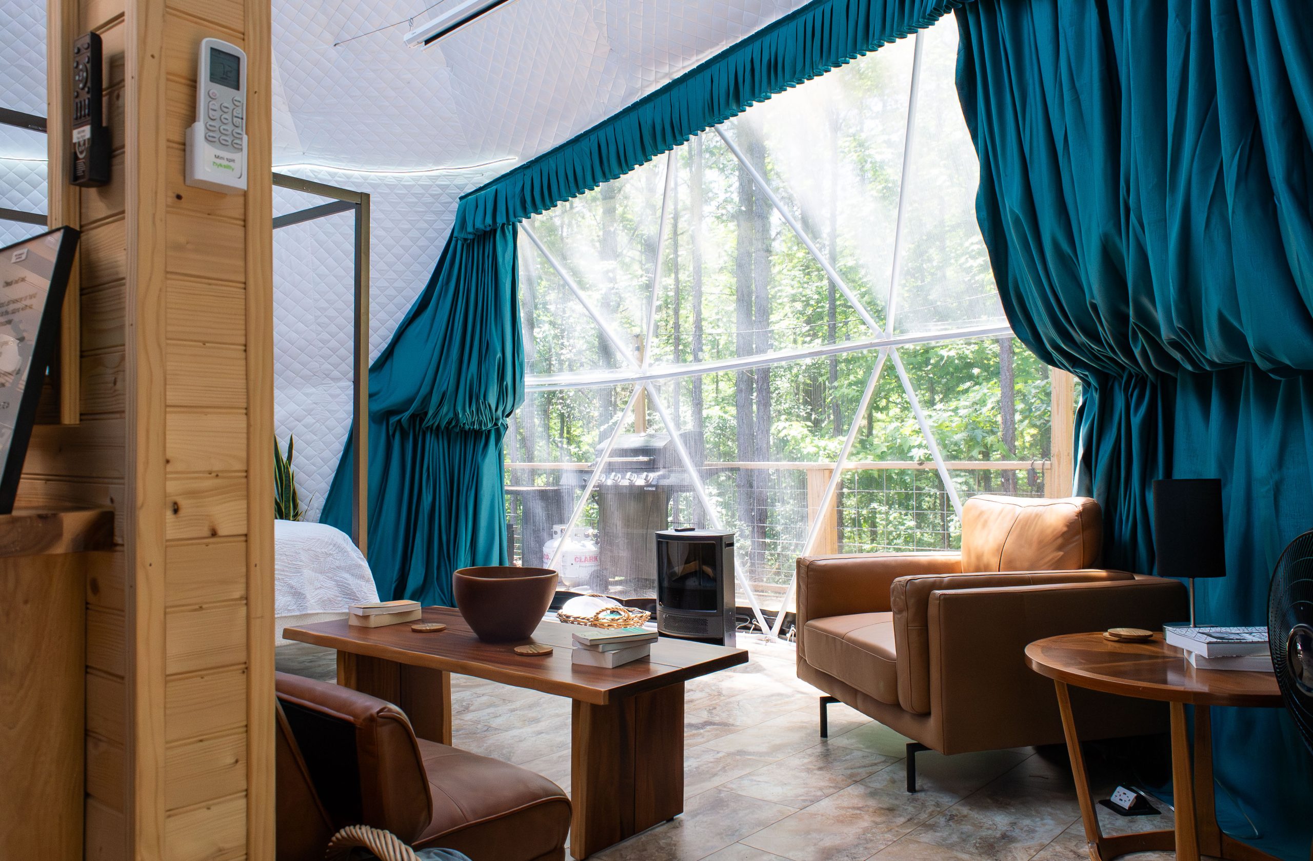 an inside image of a geodesic dome showcasing the living room looking outward through the very large window with blue drapes onto the outside porch surrounded by trees