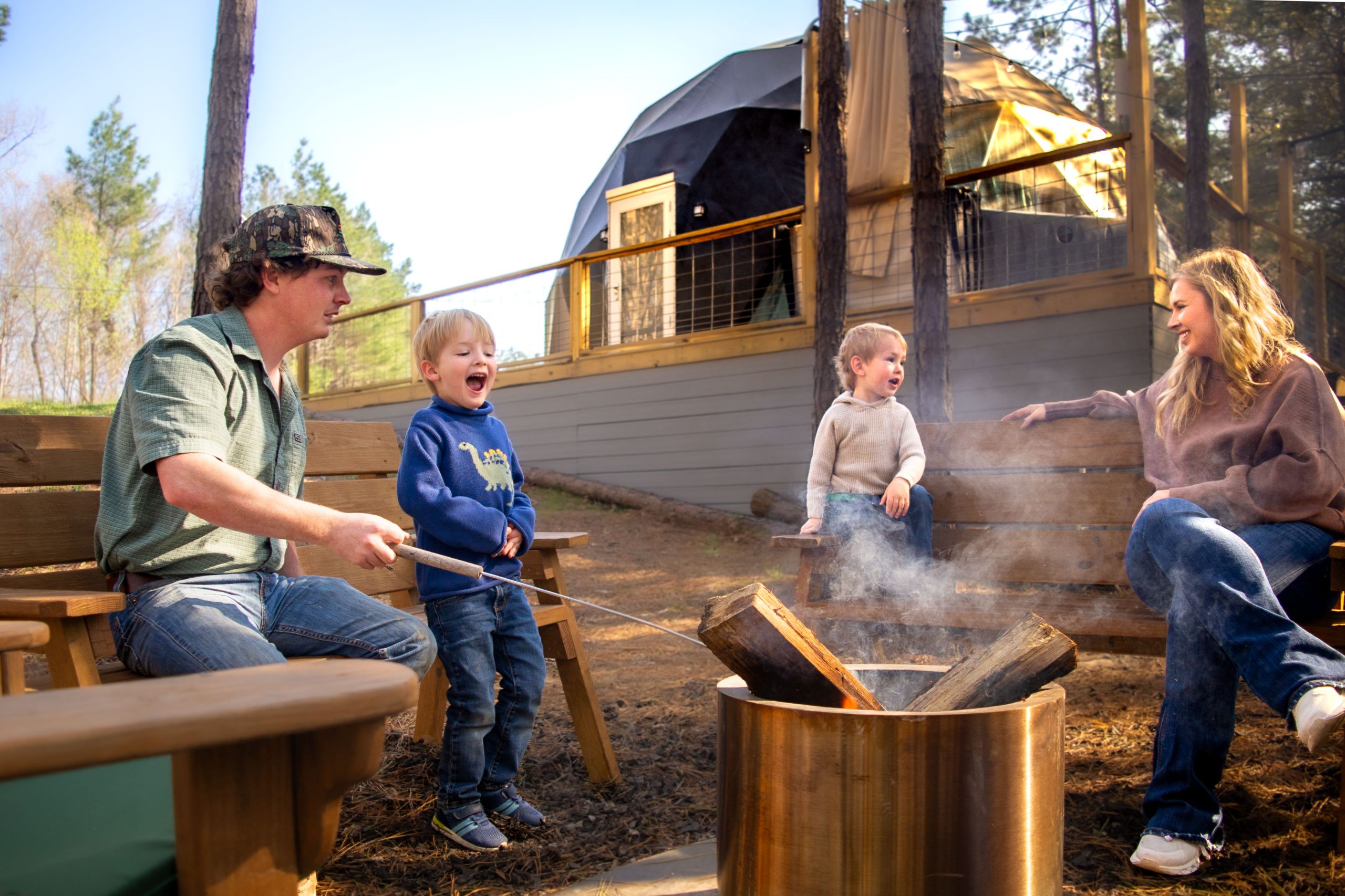 A young mother and father share marshmallows over a fire with two young boys who are laughing with a geodesic dome behind them