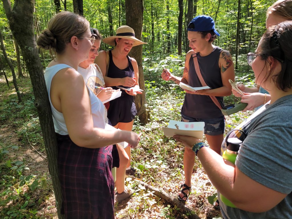 A group of ladies dressed in tank tops and hats for summer are standing in a circle holding plants and pieces of paper discussing natural pigments found outside.