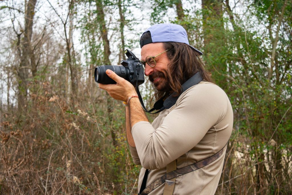 Man wearing a backwards cap is using a camera with a large lens to capture migratory birds in the evening