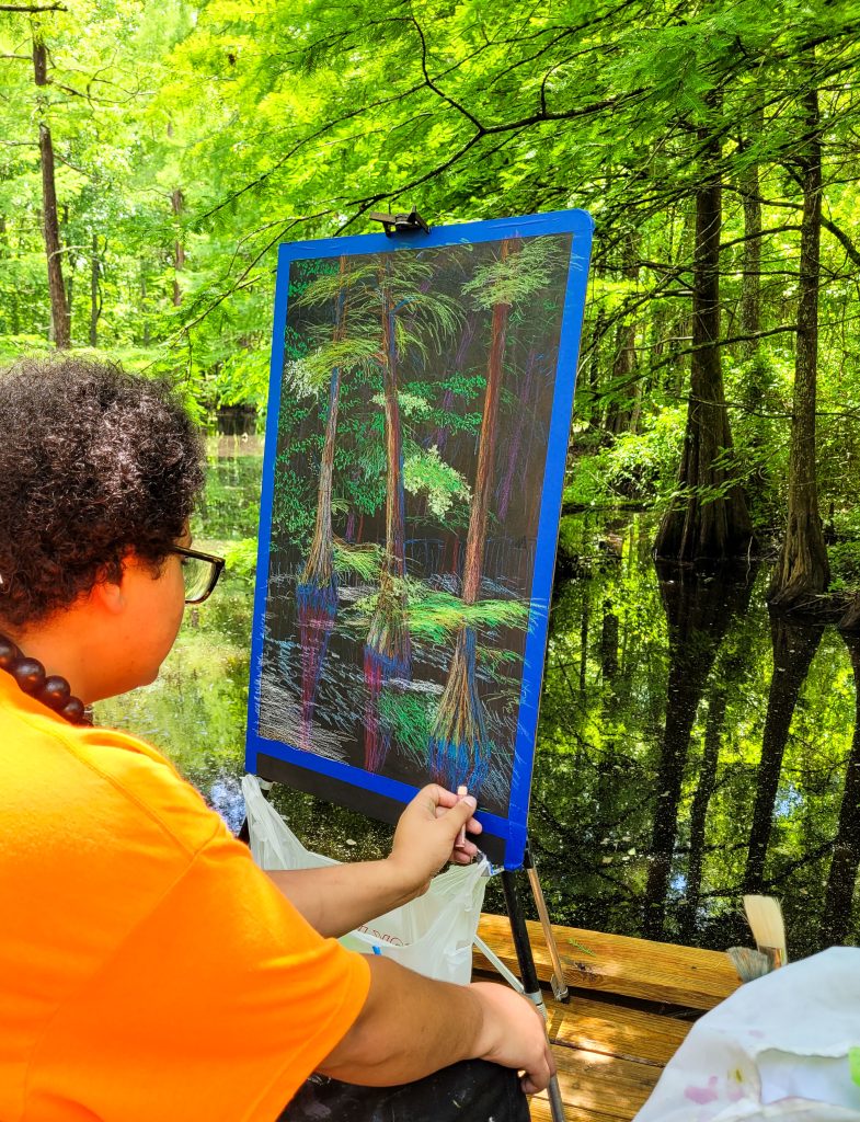 a man with an orange shirt and curly hair is using pastels to recreate the cypress trees by using an easel and black paper.