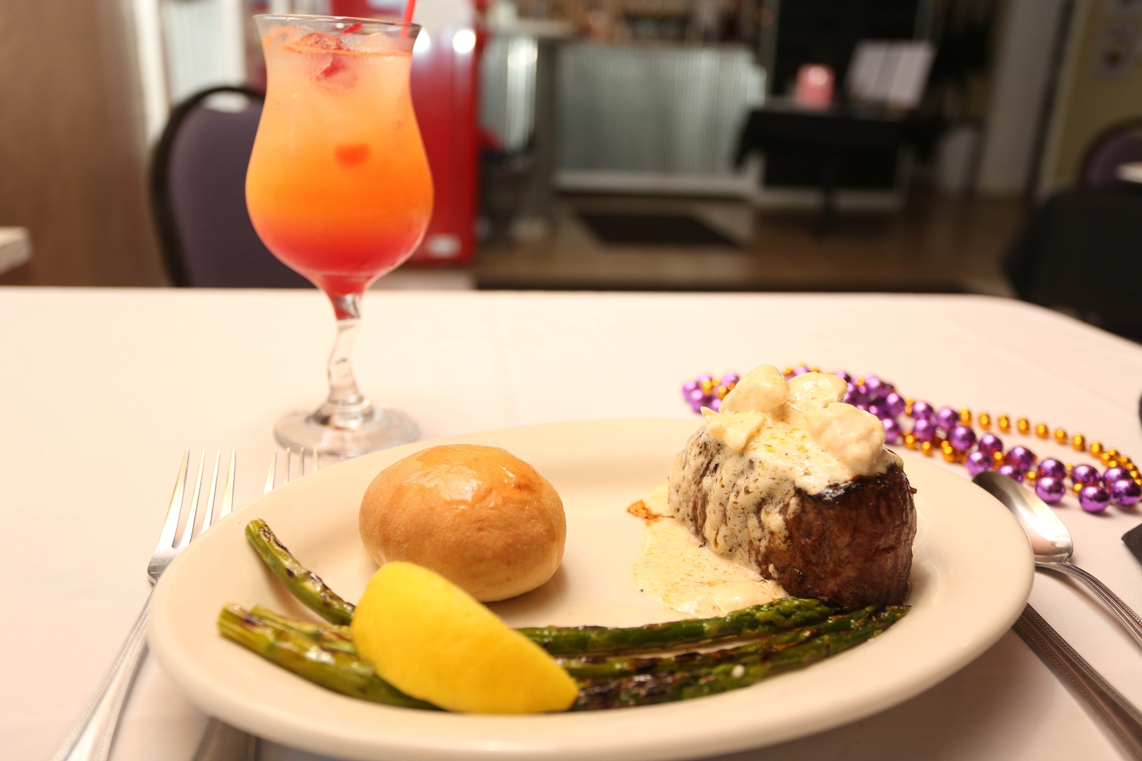 a dinner plate consists of steak with a. butter sauce, asparagus and a roll with Mardi Gras beads on the table and a mixed drink